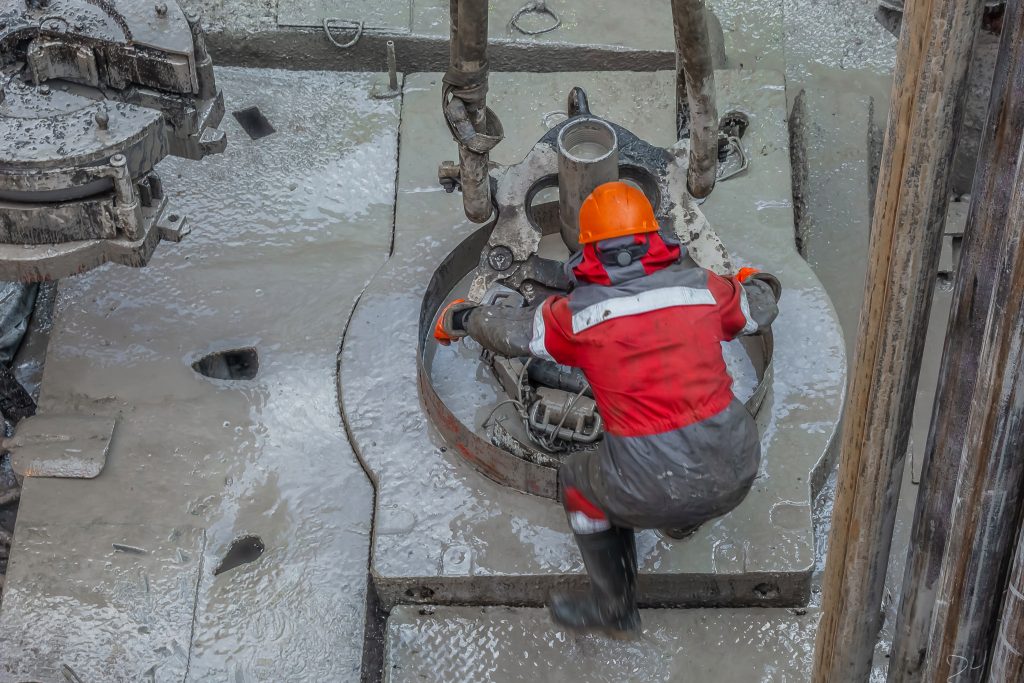 Work driller in red uniform, in helmet and goggles. He with the help of an elevator hangs drill pipes to lift them from an oil well and continue its drilling. The concept of a working person.